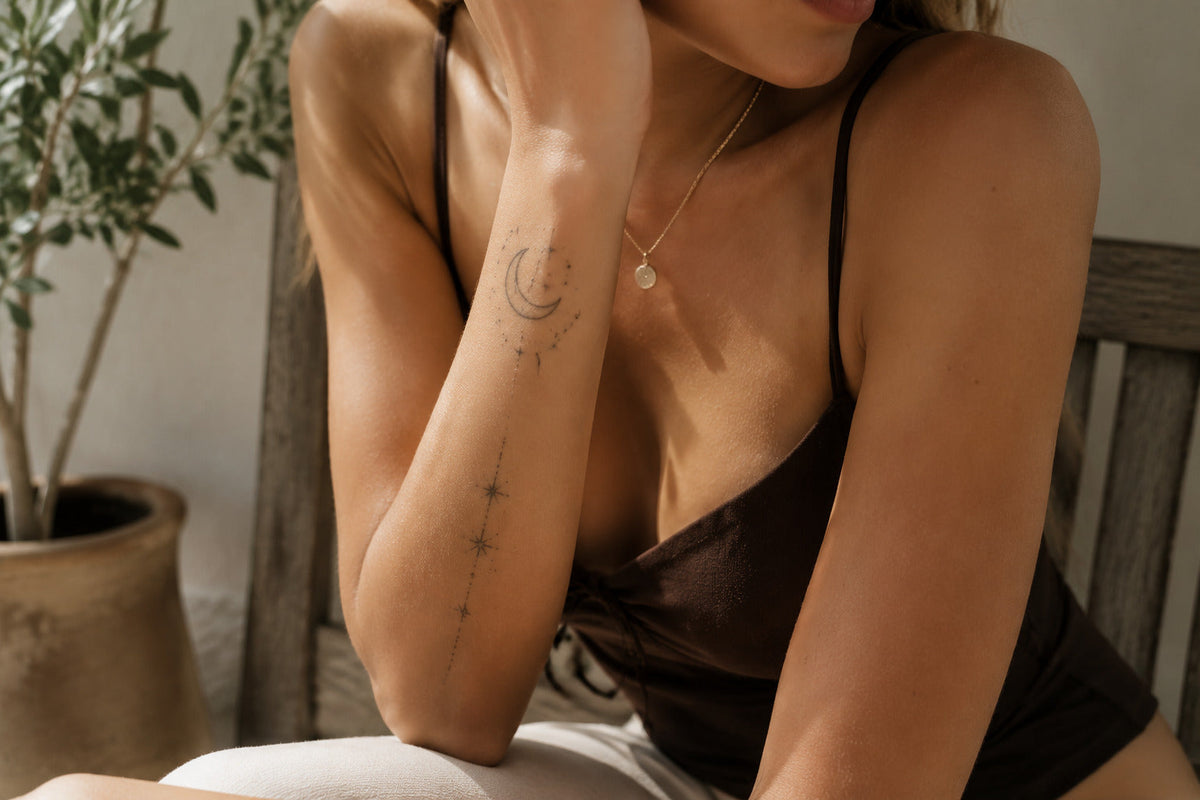 Woman wearing a crescent moon, constellation dots, and star temporary tattoo along her inner forearm in warm window light.