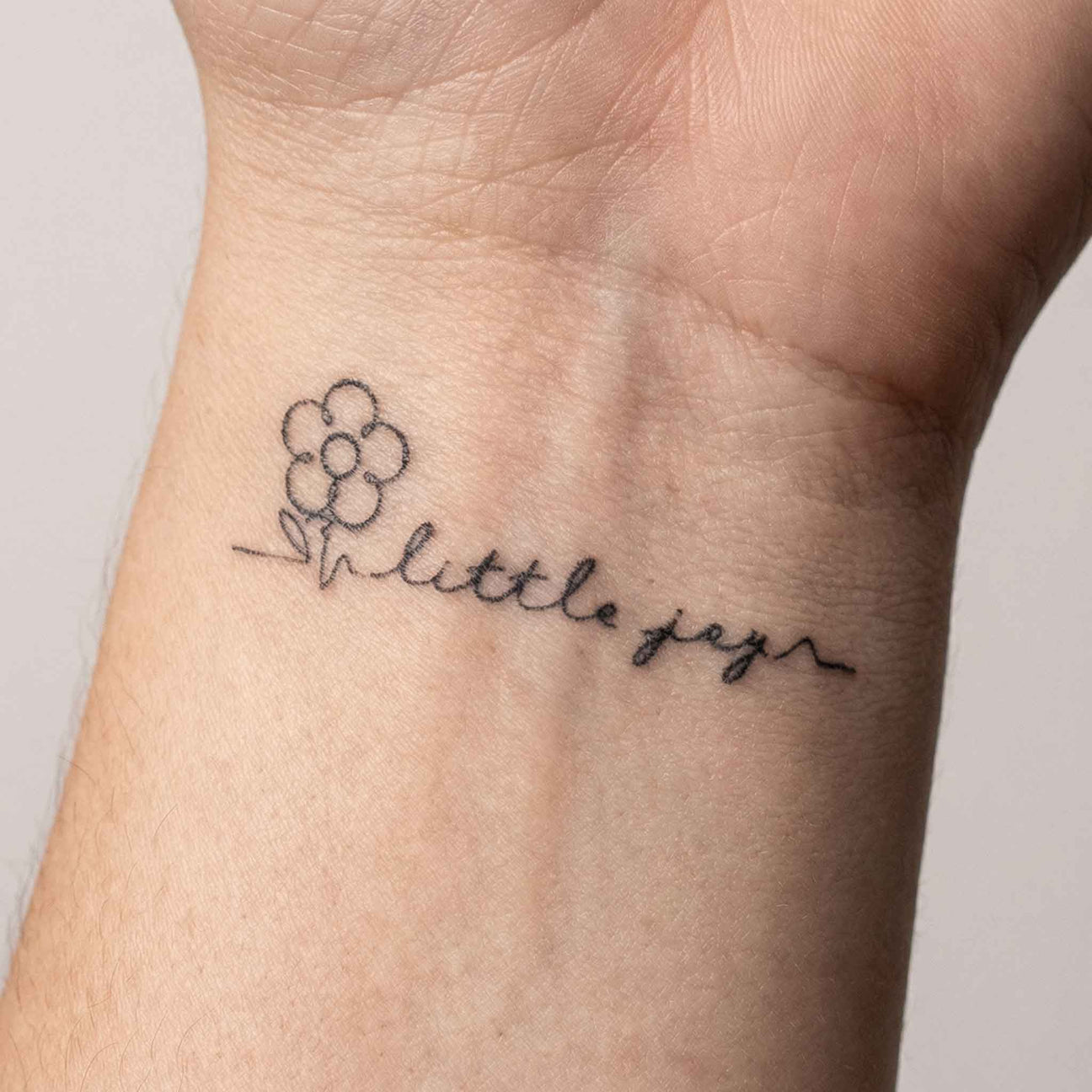 Minimal fine line semi-permanent tattoo on the wrist featuring a small flower doodle and the handwritten phrase “little you” in delicate script style.