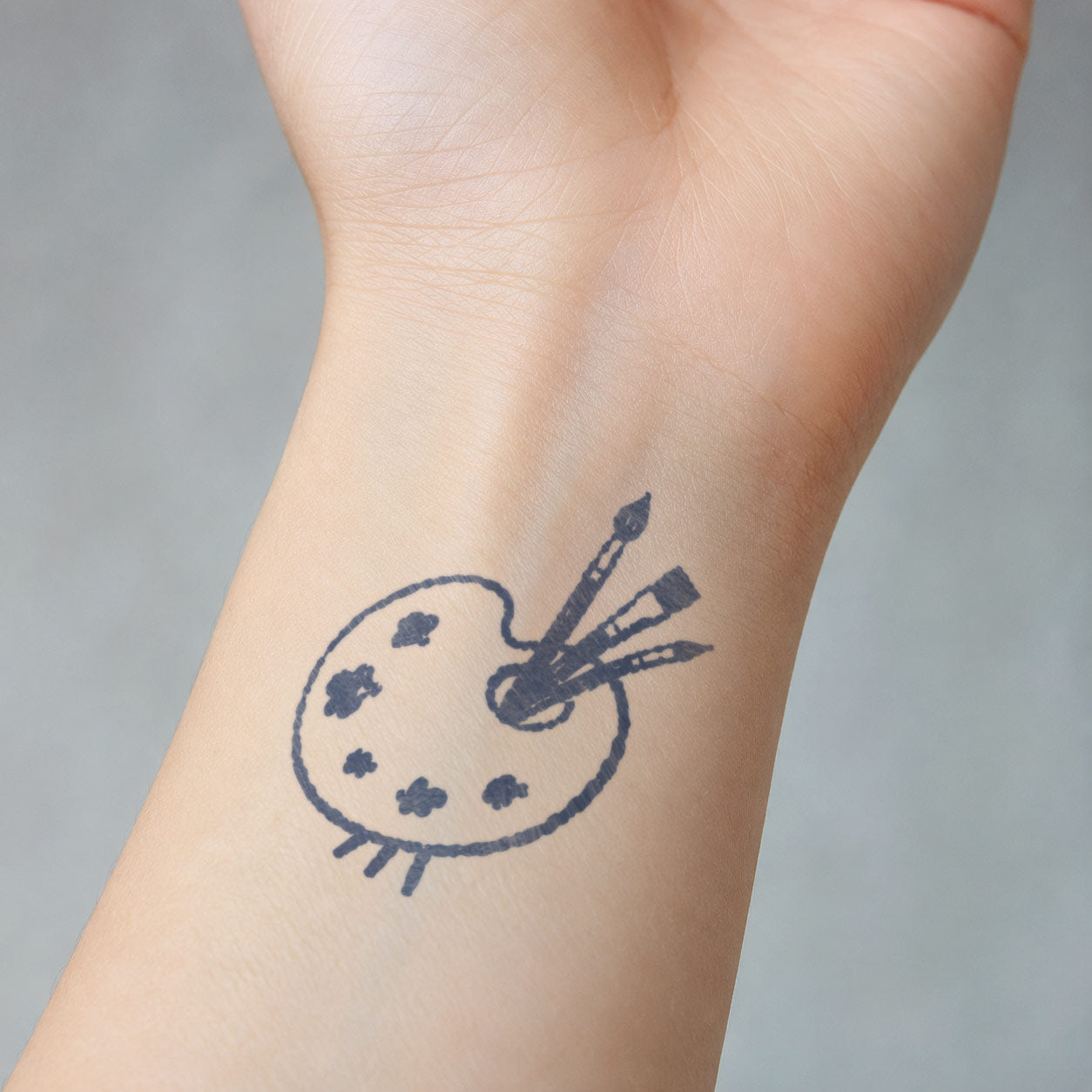 Small semi-permanent tattoo showing a painter’s palette with three brushes and scattered paint marks, placed on the inner wrist.