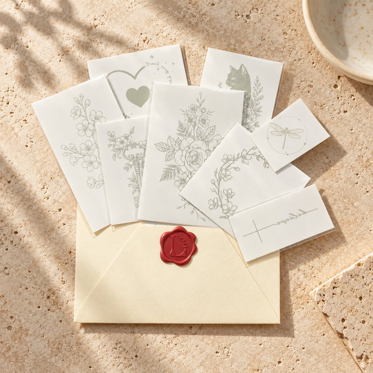 Assorted temporary tattoo sheets featuring flowers, a cat, heart, dragonfly, cross and minimalist line art arranged on a cream envelope.