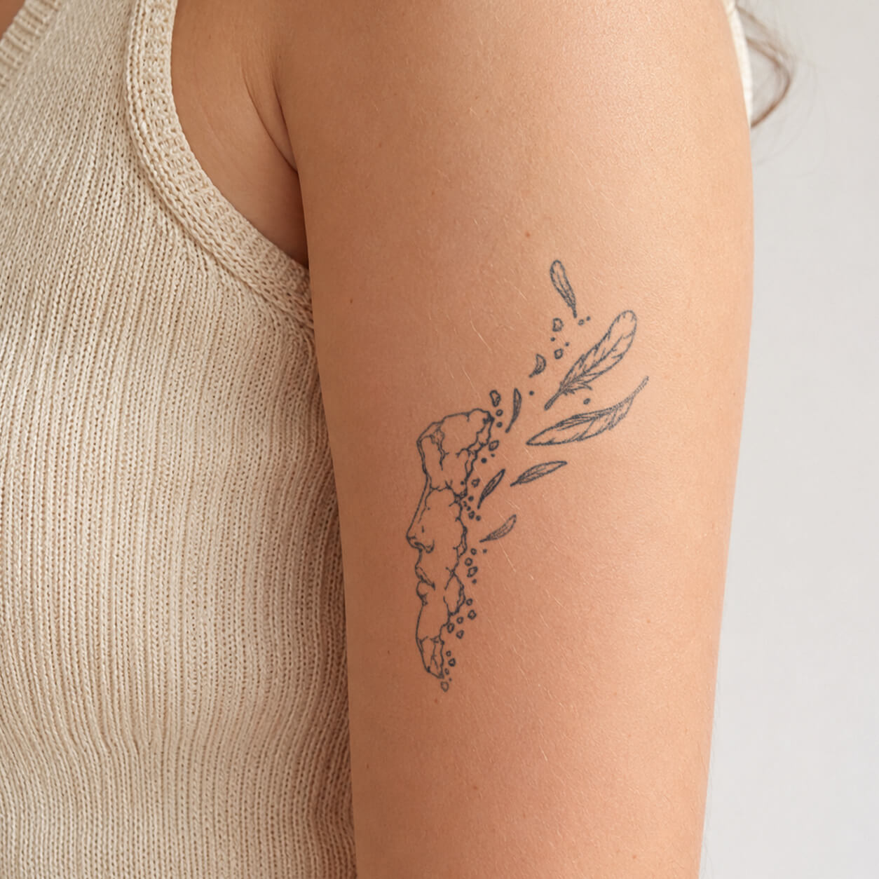 Fine line tattoo featuring a fragmented classical face sculpture with broken stone details, floating feathers, and scattered elements on the upper arm. A unique temporary tattoo inspired by ancient art, Greek sculpture, and abstract broken beauty aesthetics.