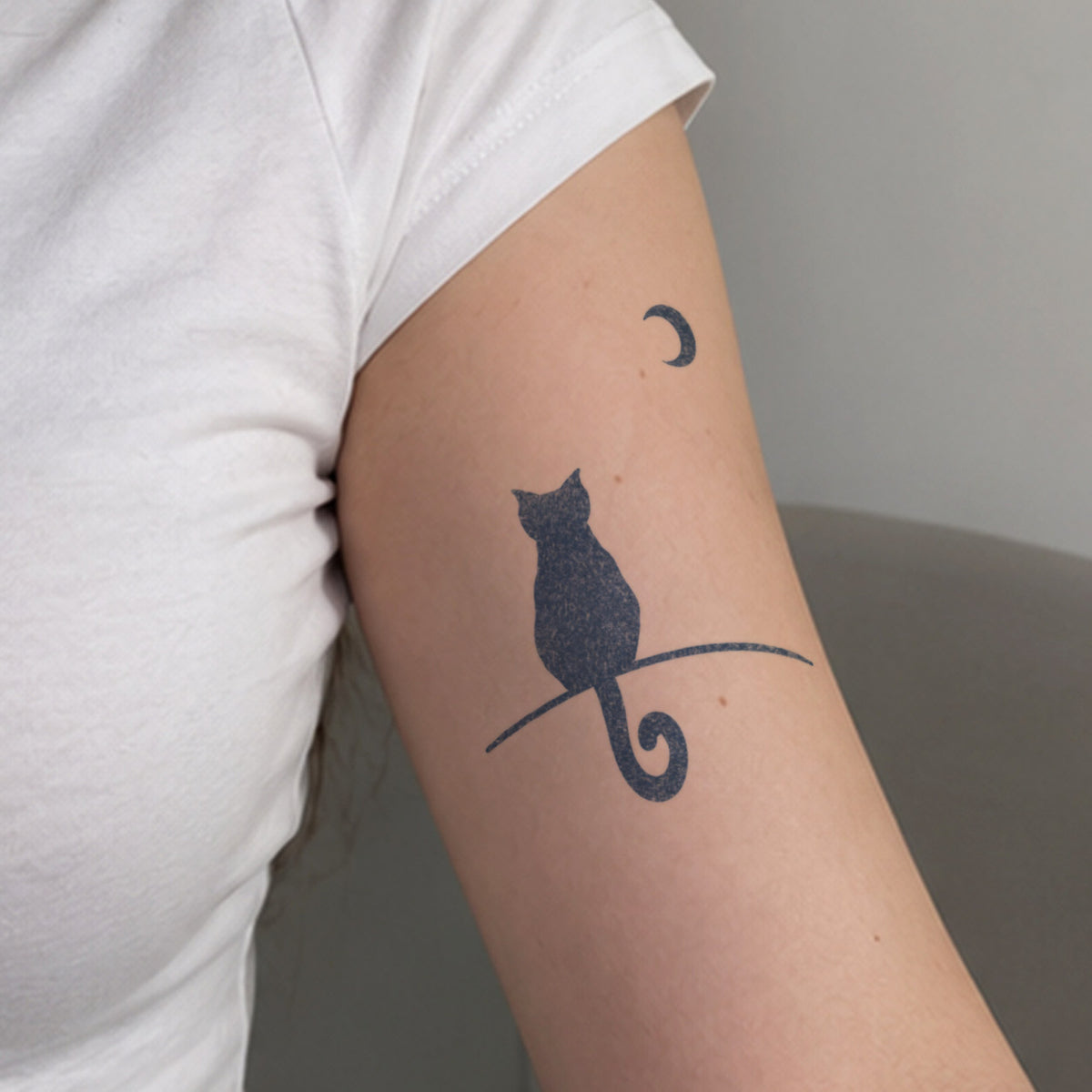 Small temporary tattoos featuring a black cat silhouette sitting beneath a crescent moon in blue-black ink on the upper arm.