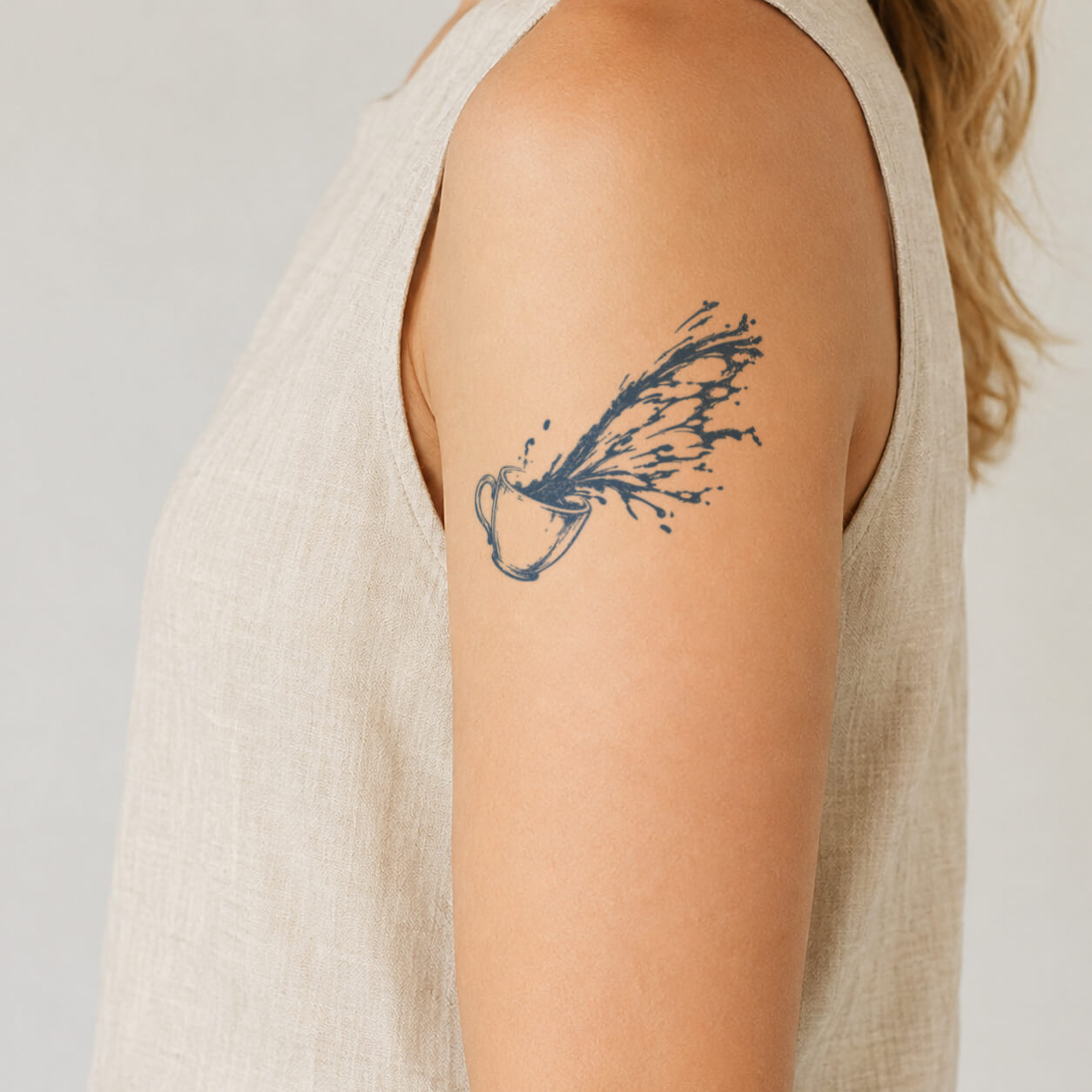 Close-up of a coffee splash tattoo featuring a small coffee cup with flowing liquid splashing upward from the rim. This creative temporary tattoo design captures the movement of coffee with artistic line details, creating a unique caffeine inspired style on the upper arm.