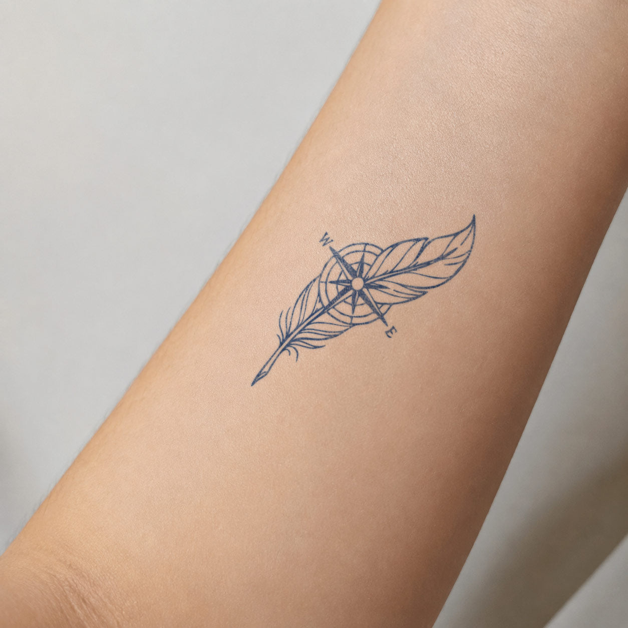 Fine-line semi-permanent tattoo featuring a feather layered with a compass on the inner forearm.