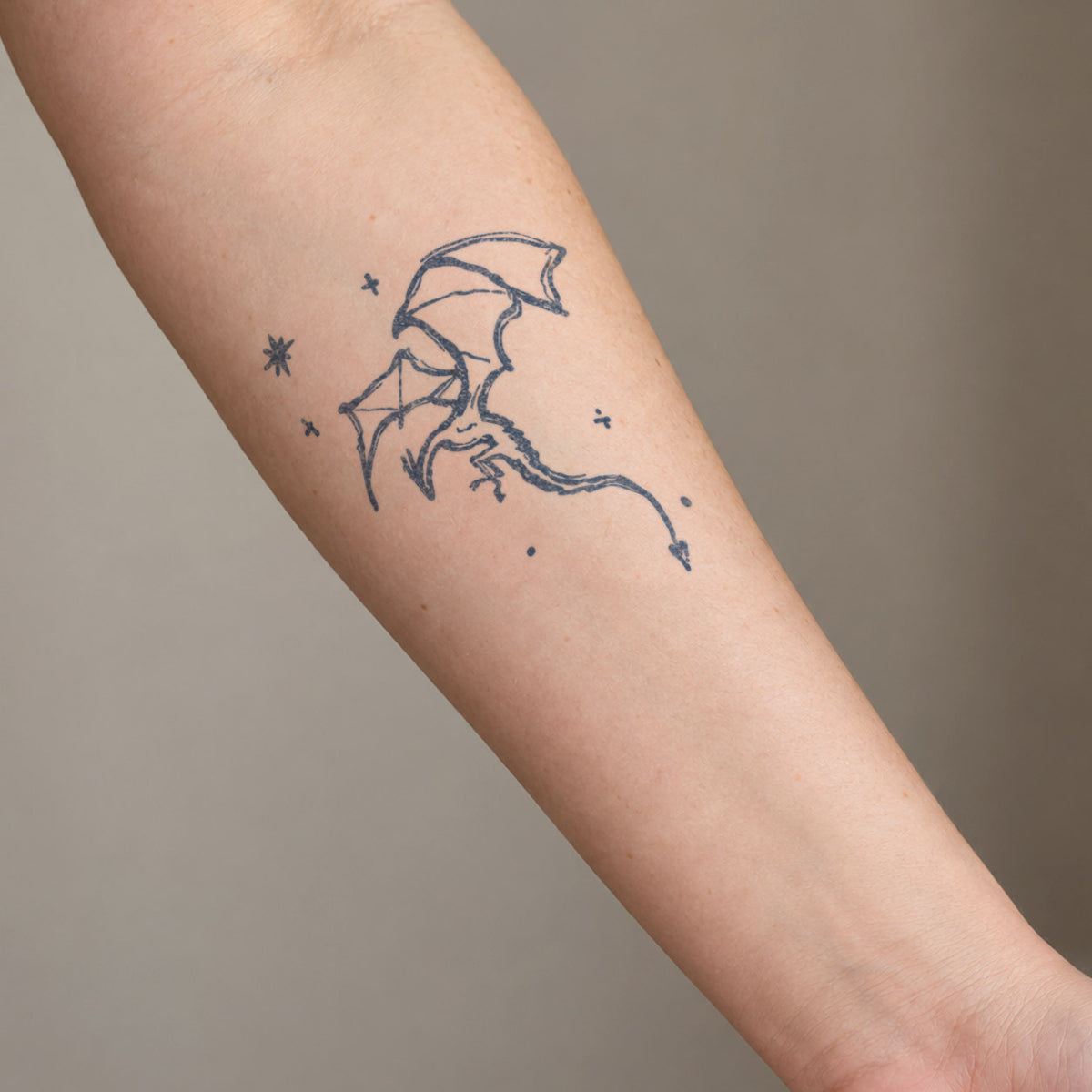 Fine line temporary tattoos featuring a flying dragon surrounded by small star details in blue-black ink on the forearm.