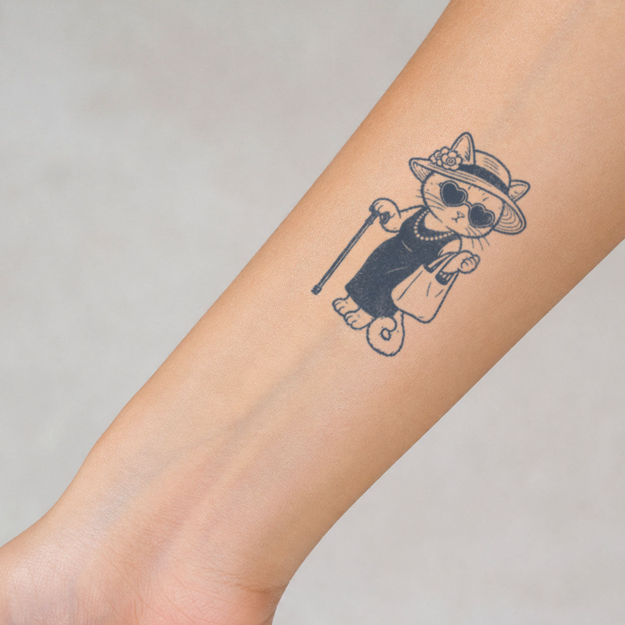 Cute temporary tattoo of a fashionable cat wearing heart-shaped sunglasses and a sun hat, shown on the forearm.