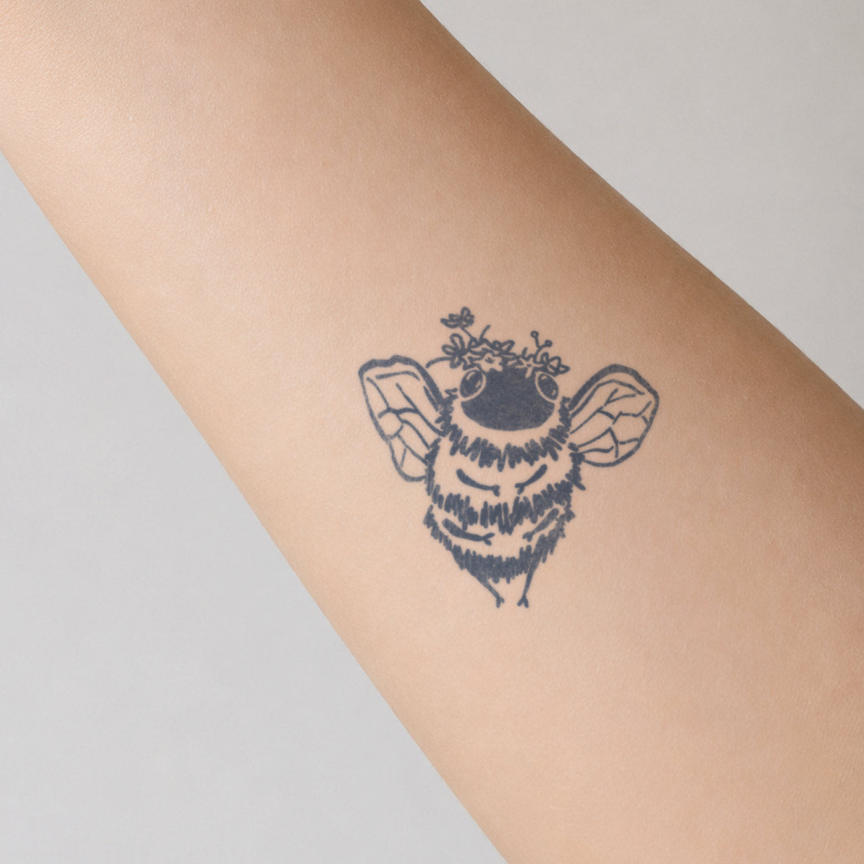 Cute semi-permanent tattoo of a fuzzy bee wearing a tiny flower crown, shown with delicate wings on the forearm.
