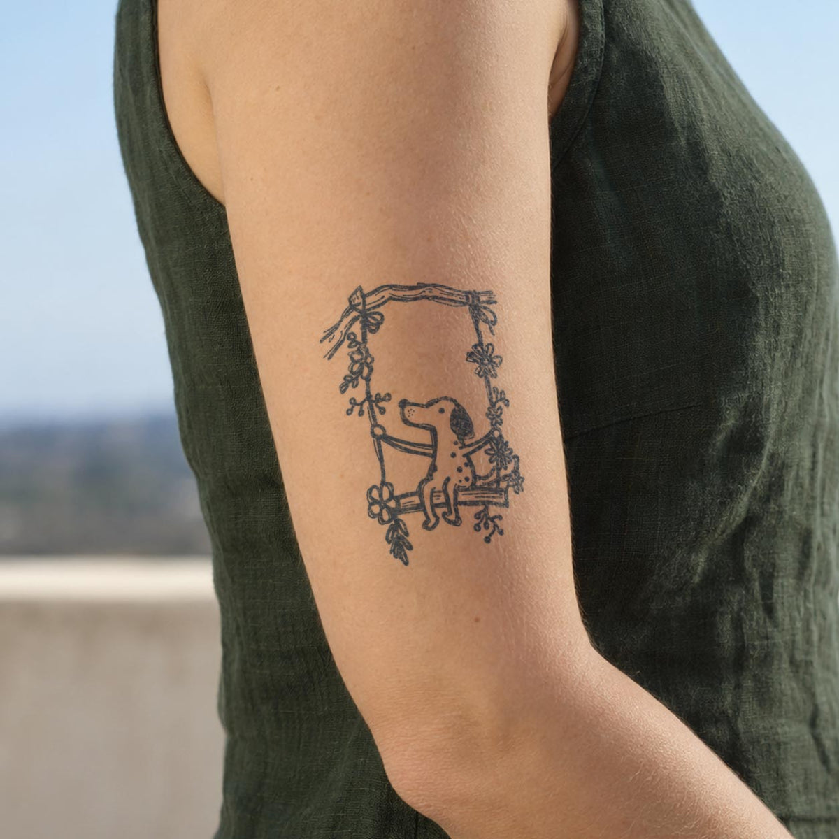 Nature temporary tattoos featuring a cute dog sitting on a flower-decorated swing in blue-black ink on the upper arm.