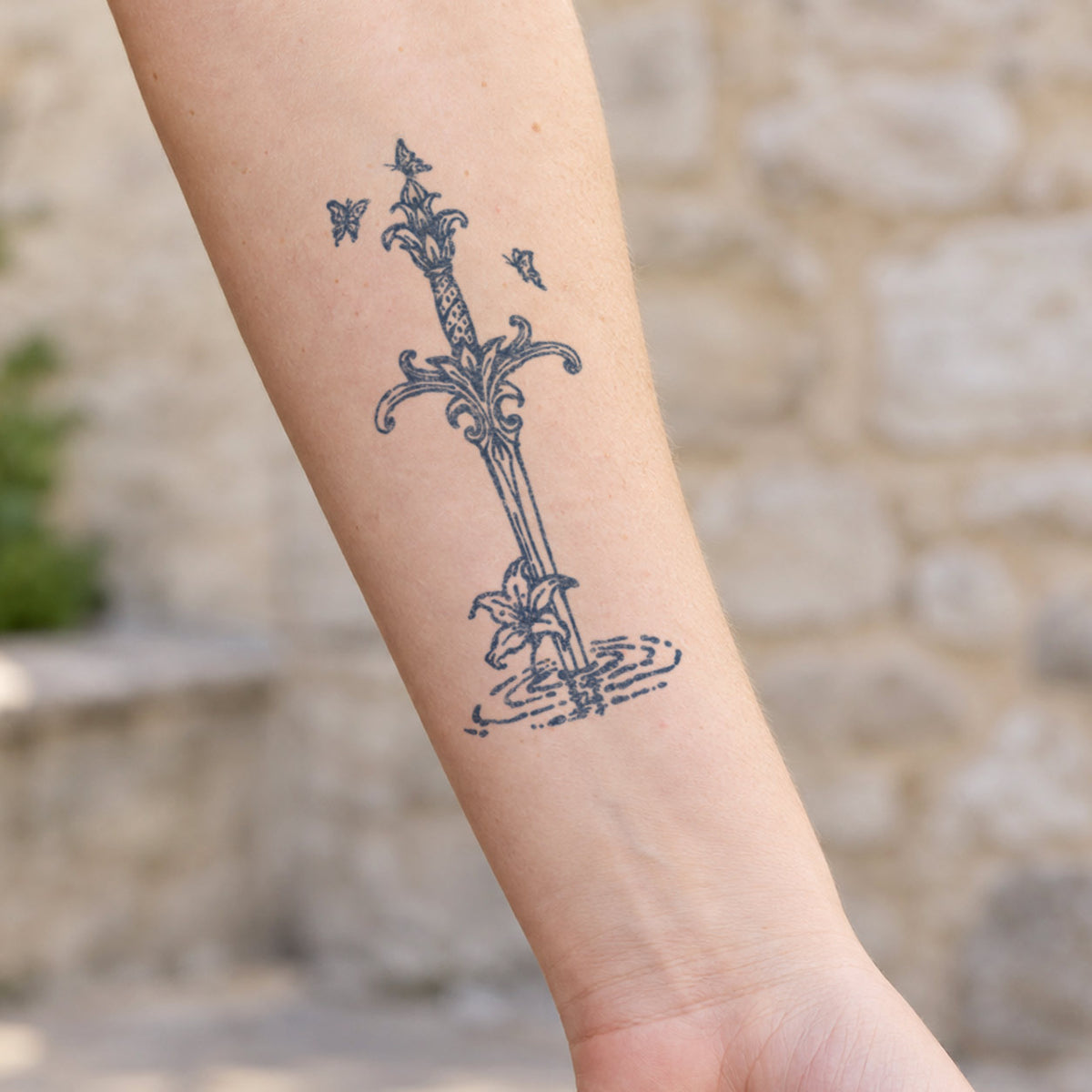 Butterfly temporary tattoos featuring an ornate sword decorated with lily flowers and small butterflies in blue-black ink on the forearm.