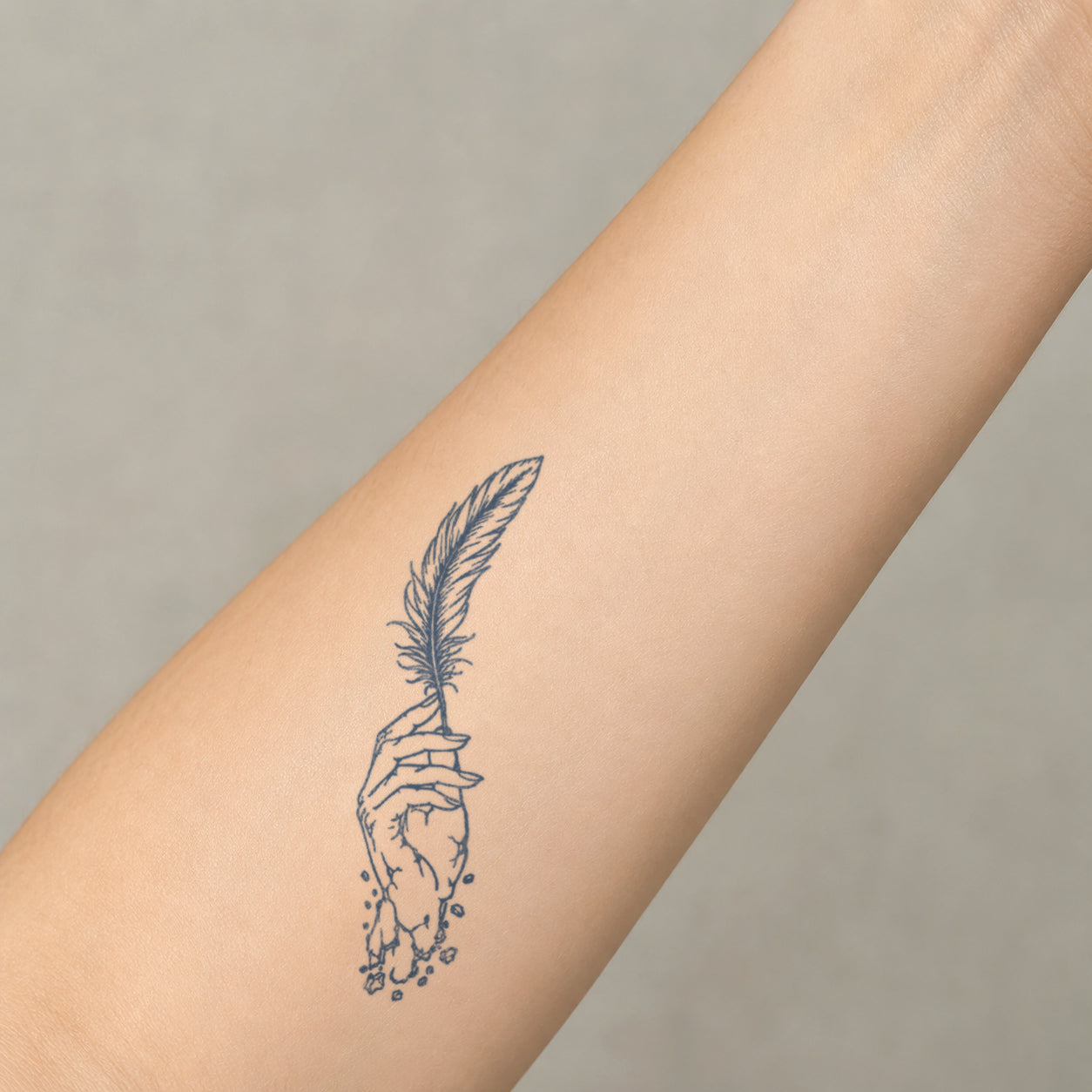 One of our detailed temporary tattoo designs featuring a fine-line hand holding a delicate feather, displayed vertically on the inner forearm.