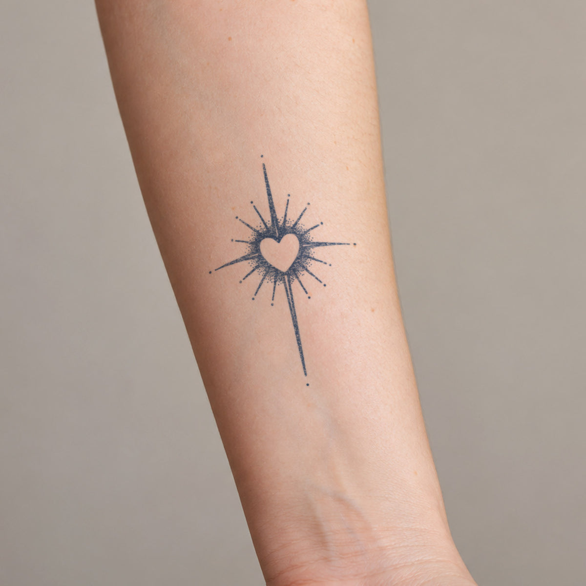 Minimalist temporary tattoos featuring a small heart surrounded by fine radiating lines in blue-black ink on the inner forearm.
