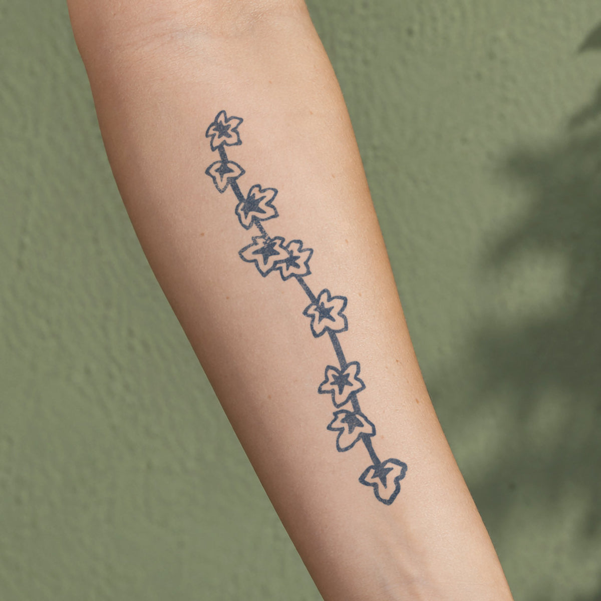 Nature temporary tattoos featuring a minimalist ivy vine with small leaves in blue-black ink along the forearm.