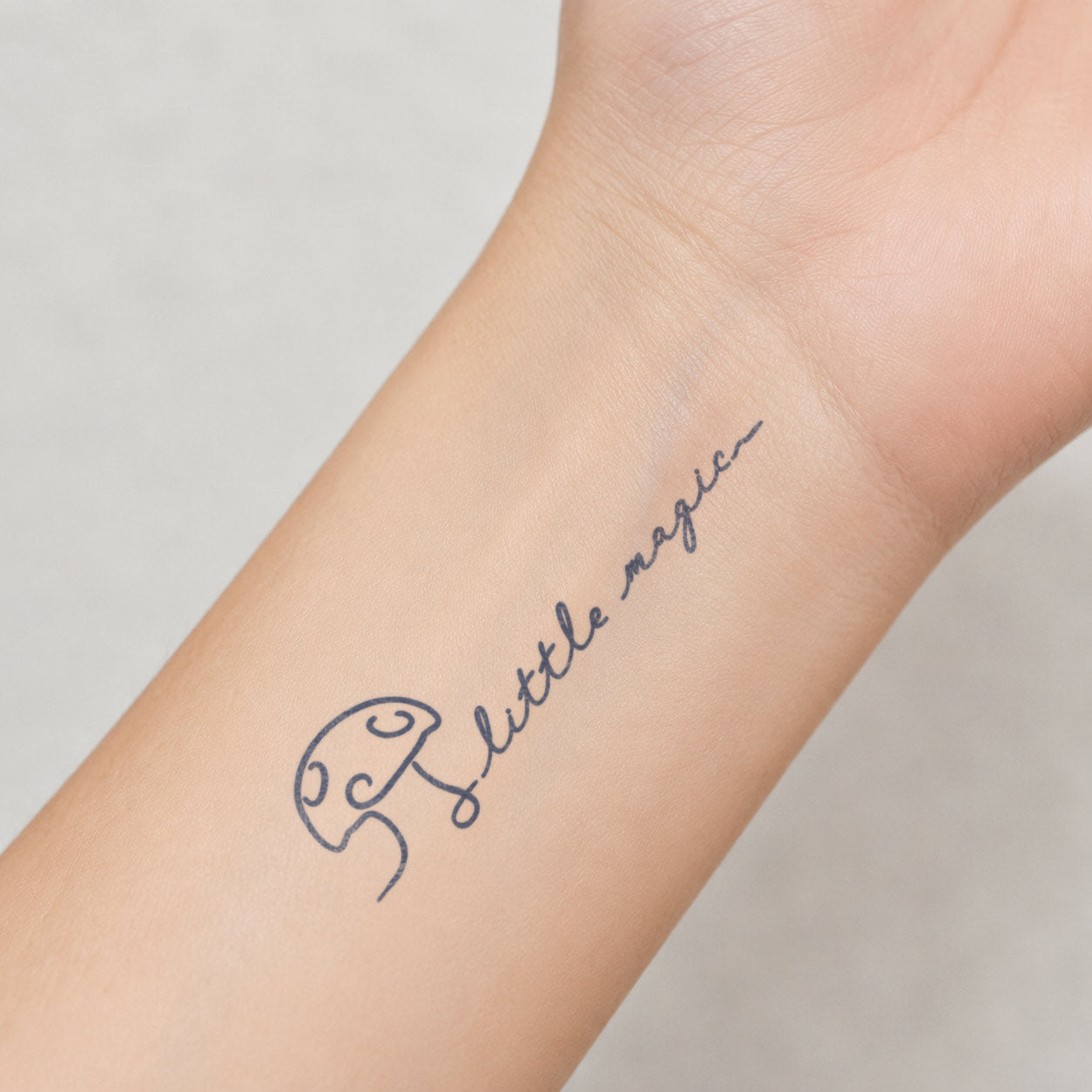 Semi-permanent tattoo featuring a Little Magic mushroom design with clean line art, tiny sparkle details, and soft handwritten lettering on the inner wrist.