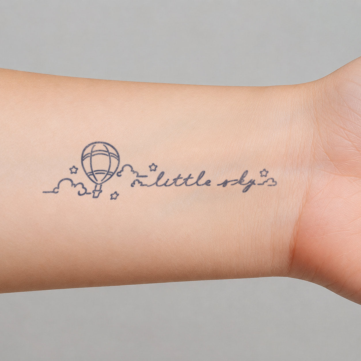 Temporary tattoo design featuring a Little Sky hot air balloon with clouds, tiny stars, and soft handwritten lettering on the inner wrist.