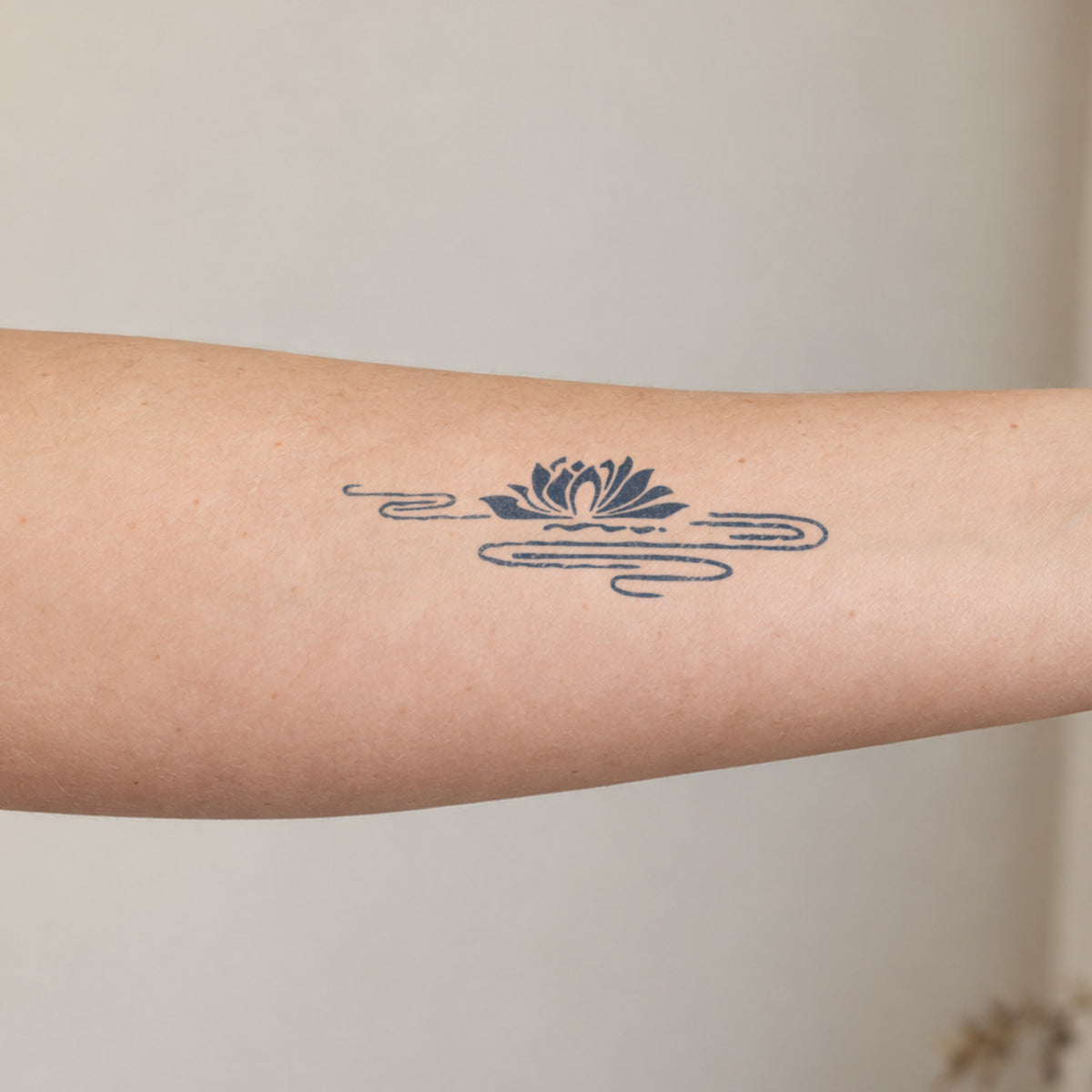 Fine line temporary tattoos featuring a minimalist lotus flower above flowing water lines in blue-black ink on the forearm.