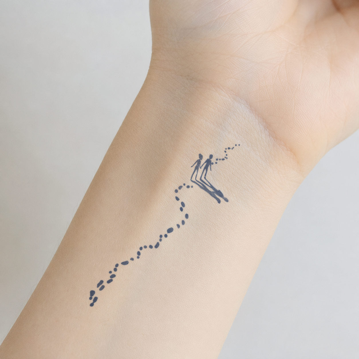 Small temporary tattoo design showing two minimal figures following a dotted path, creating a sweet adventure-inspired look on the inner wrist.