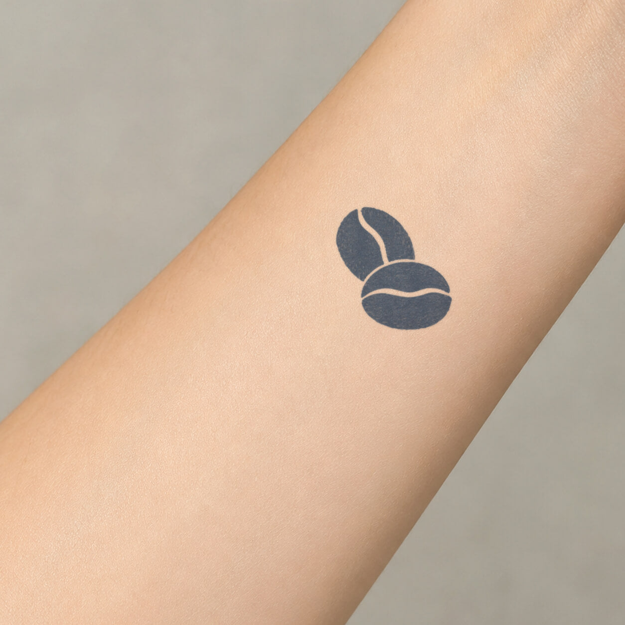 Minimal black coffee bean semi permanent tattoo placed on the inner forearm, featuring two rounded beans with clean negative-space line details.