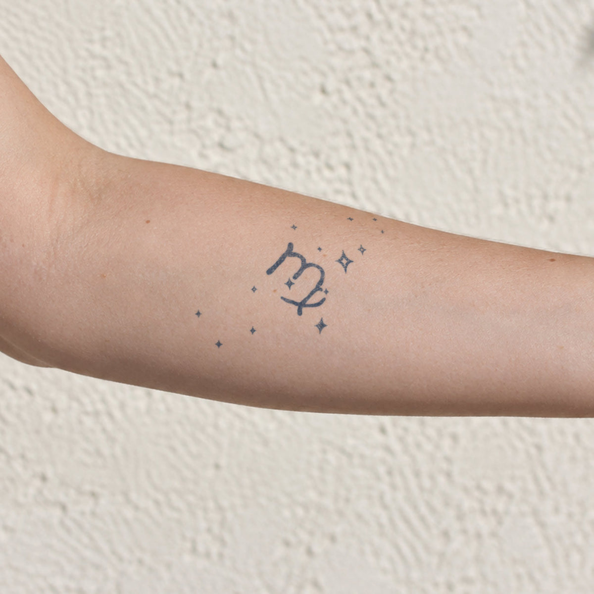 Minimalist temporary tattoos featuring a Virgo zodiac symbol surrounded by small sparkling stars in blue-black ink on the inner forearm.