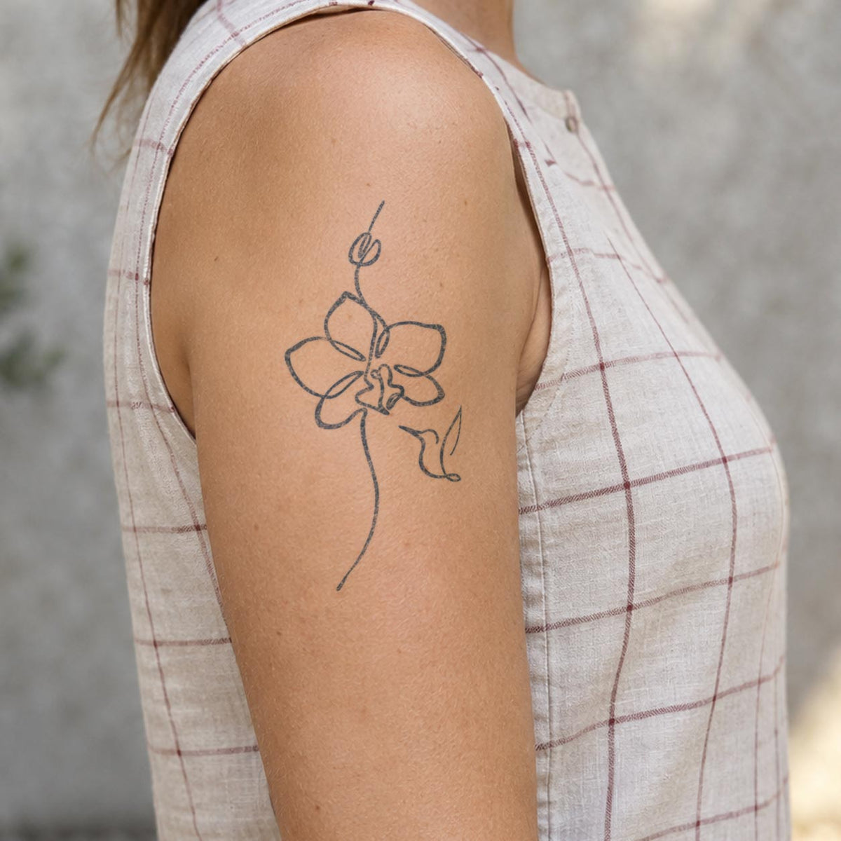 Floral temporary tattoos featuring a minimalist orchid flower with a small hummingbird in blue-black line art on the upper arm.