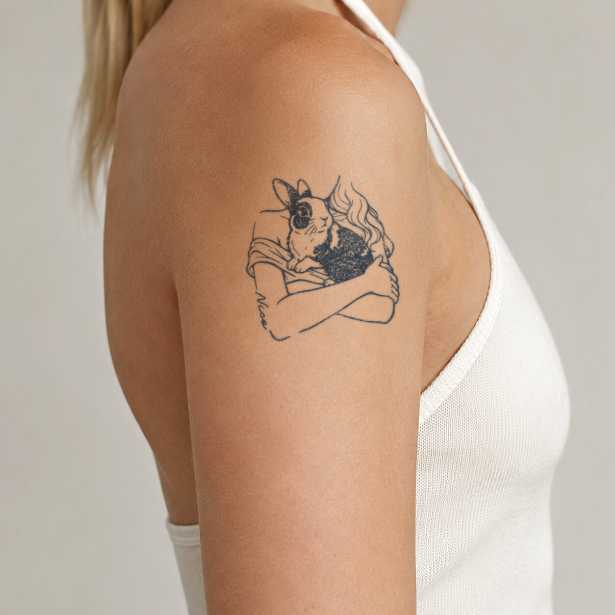 Close-up of a fine line rabbit tattoo featuring a person gently holding a bunny with flowing hair details and a handwritten name element. This sentimental pet inspired tattoo design creates a warm and meaningful look on the upper arm.