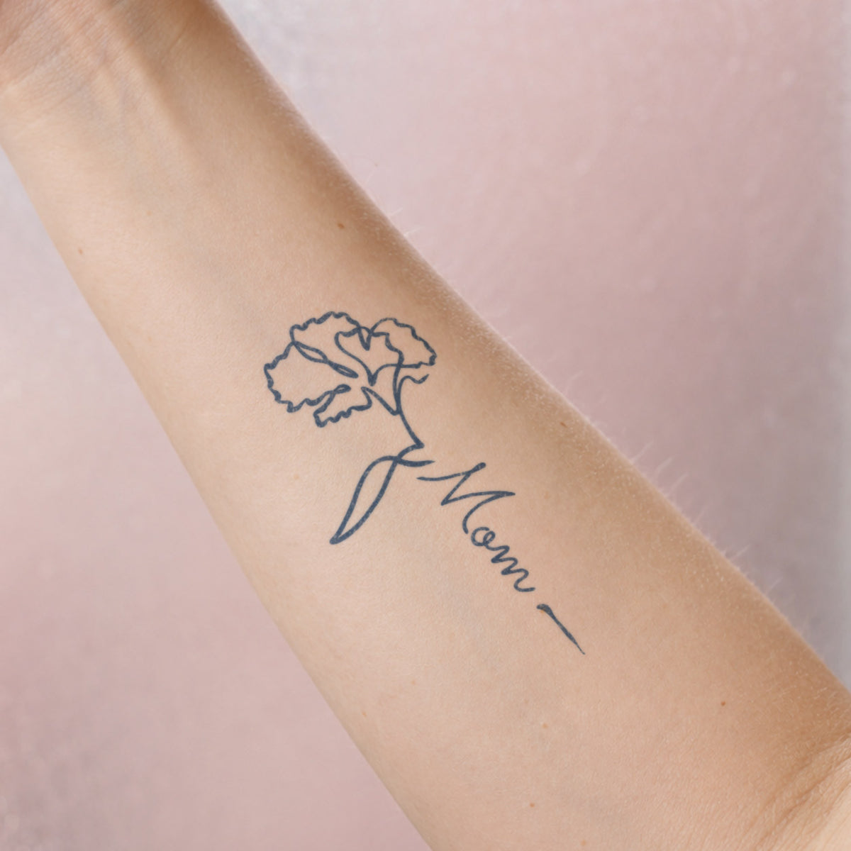 Small flower temporary tattoos featuring a delicate floral line design connected to the word “Mom” in blue-black ink on the forearm.