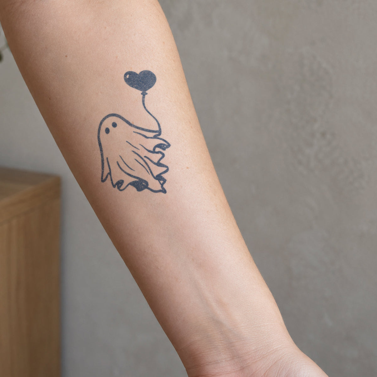 Minimalist temporary tattoos featuring a cute ghost holding a heart-shaped balloon in blue-black ink on the forearm.