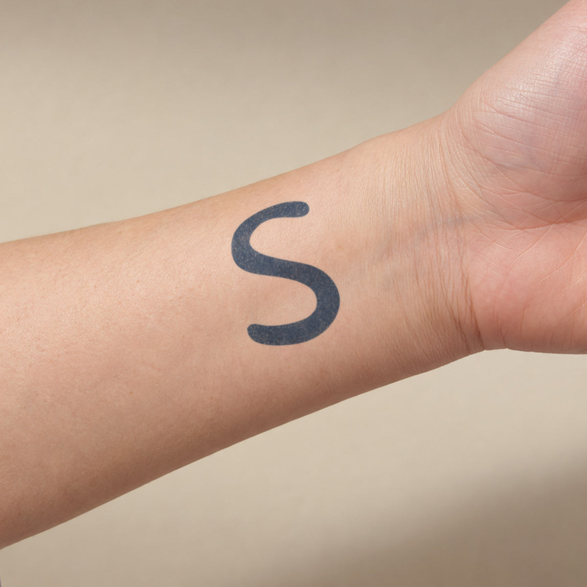 Small temporary tattoos featuring a bold minimalist S letter in blue-black ink on the inner wrist.
