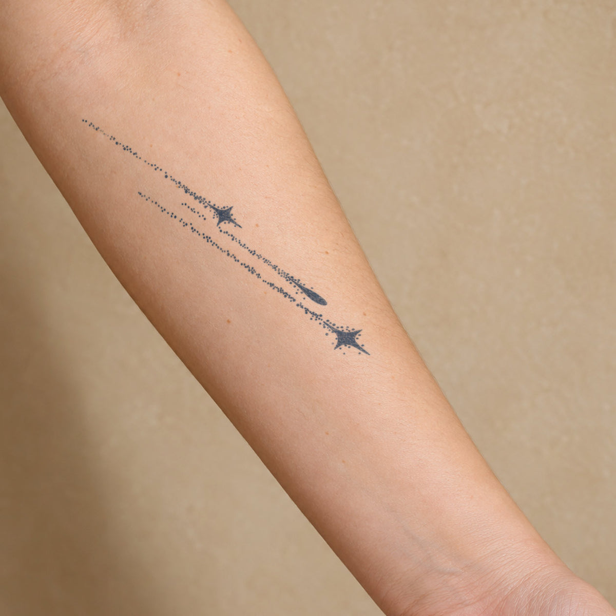 Minimalist temporary tattoos featuring shooting stars and delicate dotted trails in blue-black ink on the forearm.