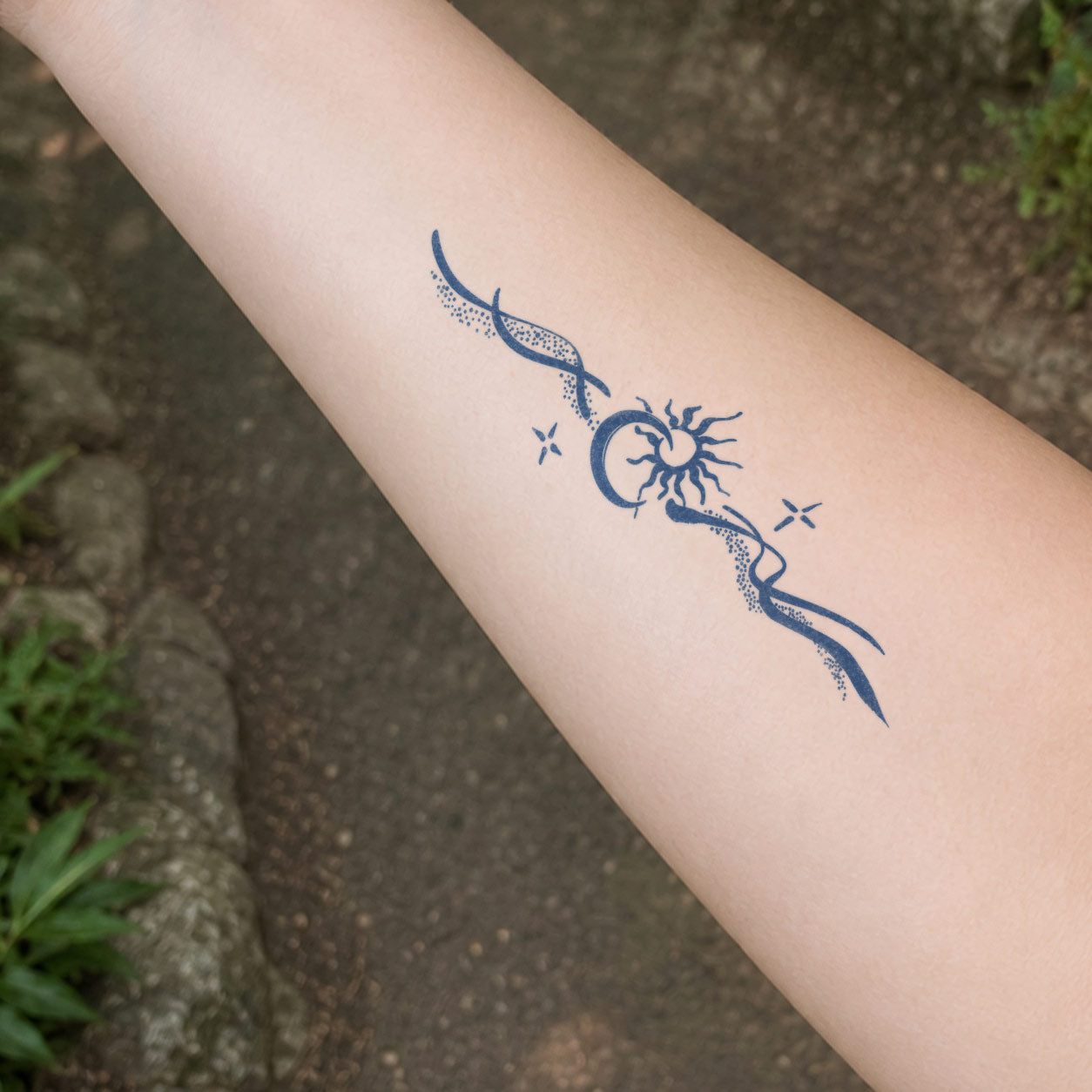 Temporary forearm tattoos featuring a crescent moon framing a radiant sun, flowing dotted ribbons, and small sparkle accents.
