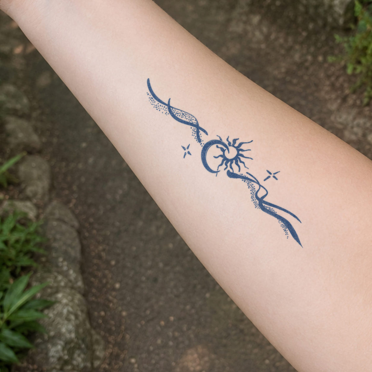 Temporary forearm tattoos featuring a crescent moon framing a radiant sun, flowing dotted ribbons, and small sparkle accents.
