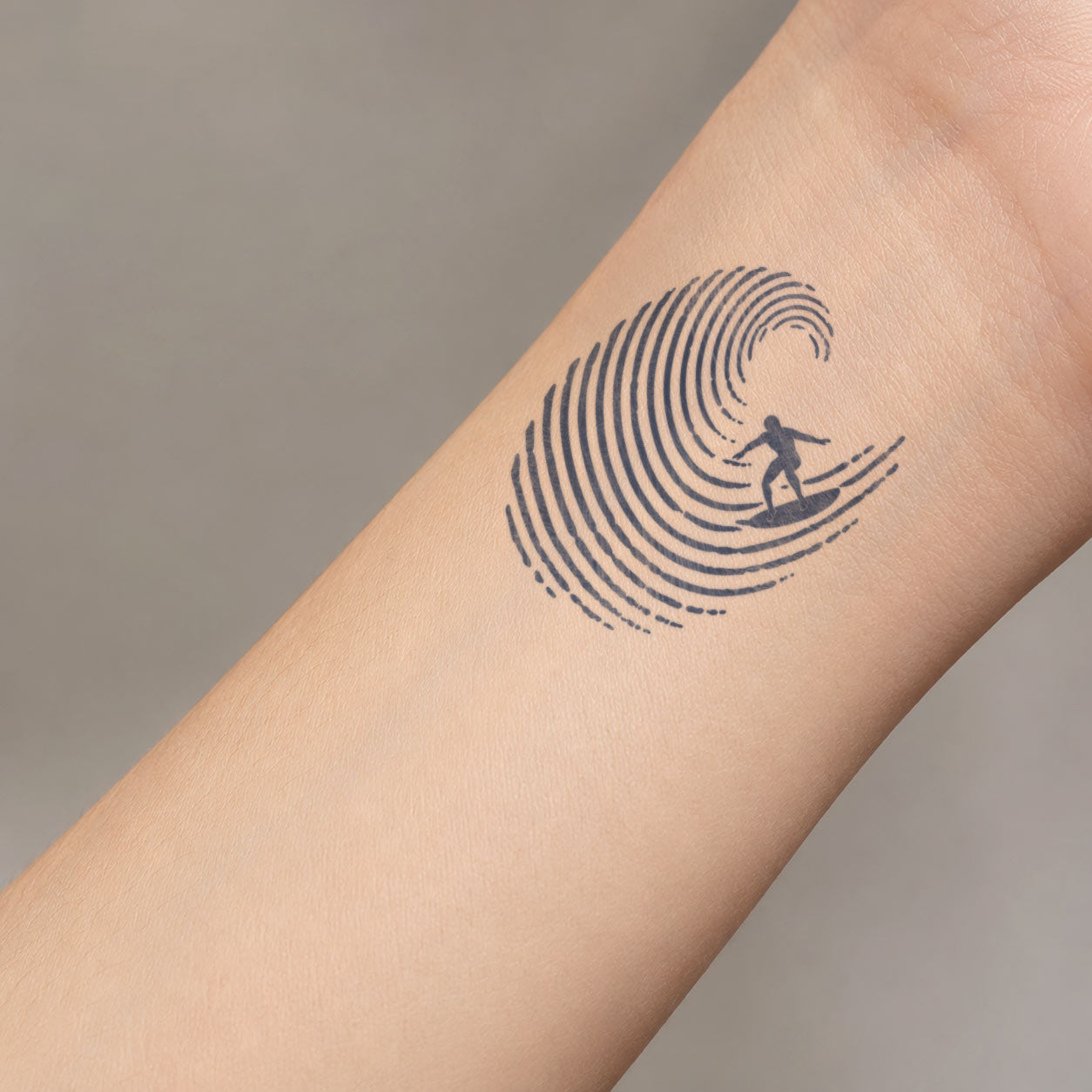 Minimal temporary tattoo design featuring a small surfer riding inside a curling ocean wave, placed on the inner forearm near the wrist.