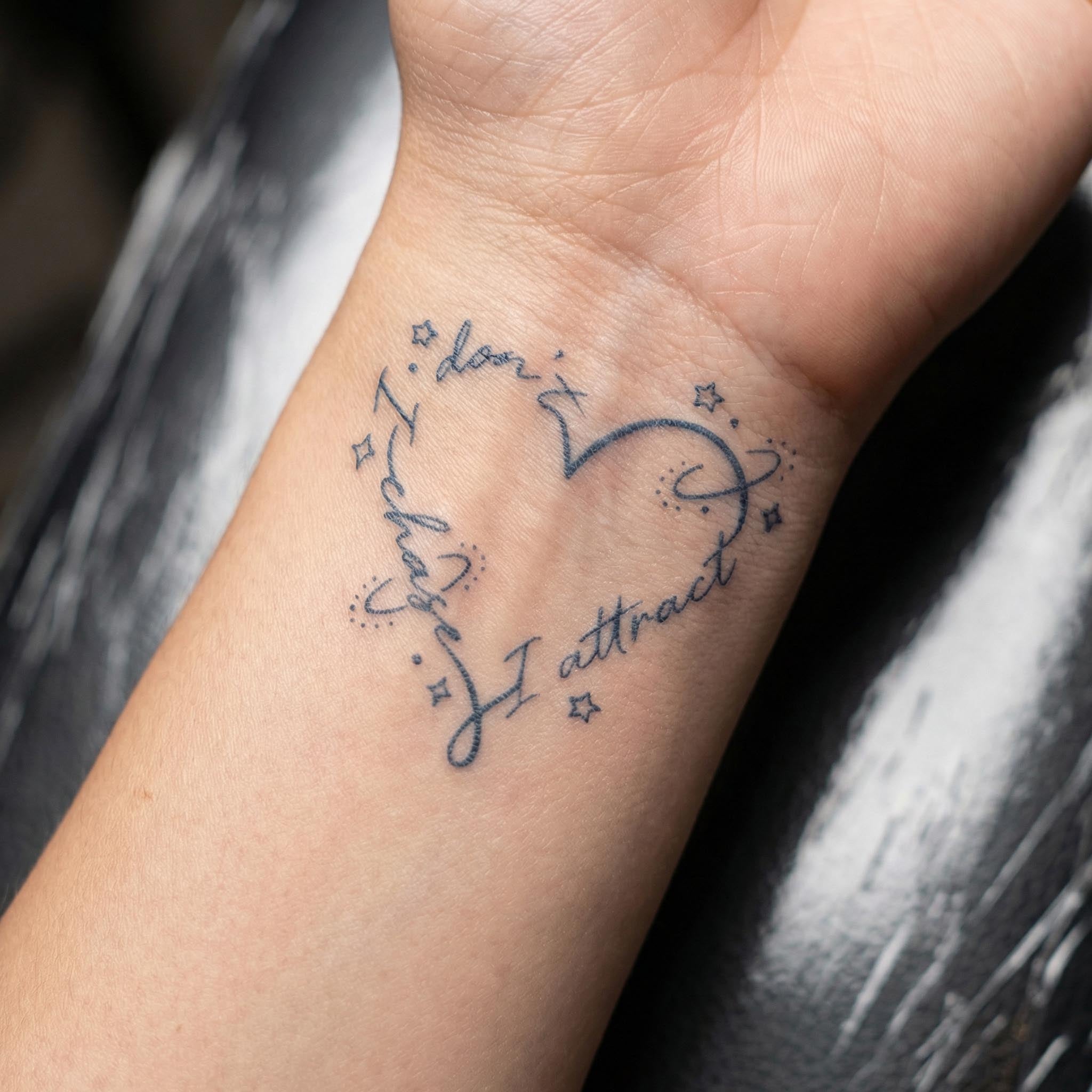 A close-up photograph of a delicate black ink tattoo on a person's inner wrist. The tattoo replicates the heart-shaped design made of the words "I don't chase, I attract" with surrounding star and celestial accents.