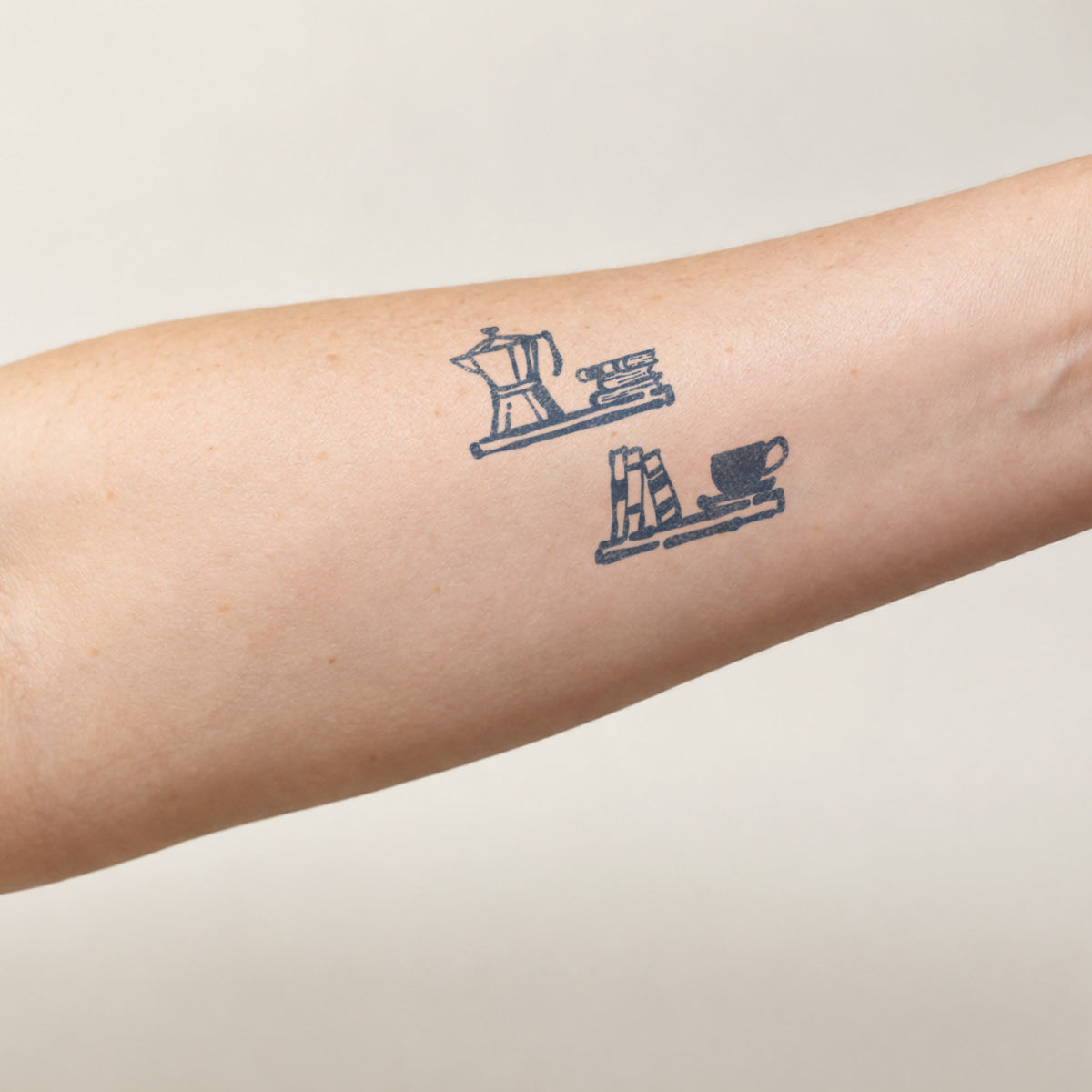 Temporary tattoo designs featuring a moka pot, stacked books, and a coffee cup in blue-black line art on the forearm.
