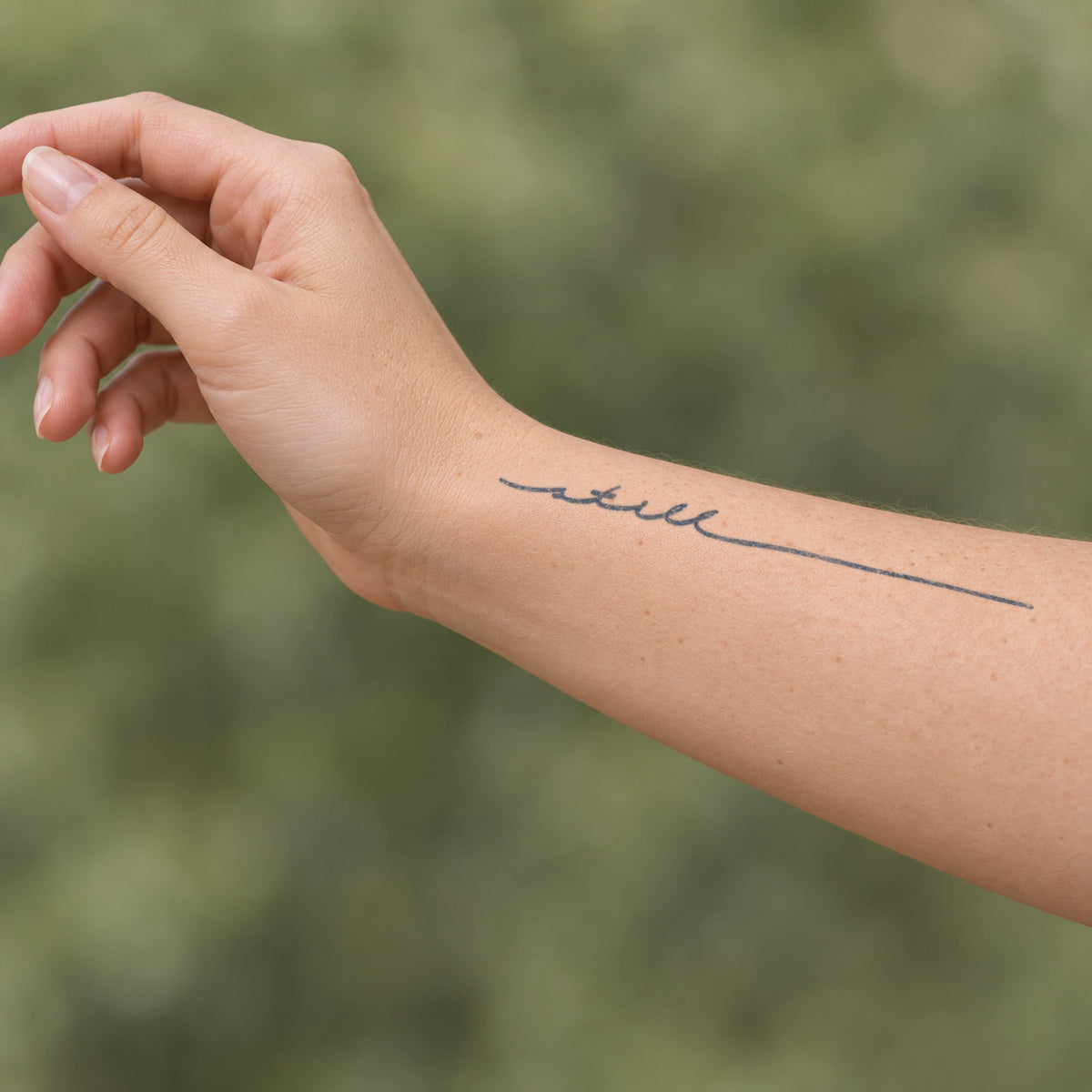 Temporary forearm tattoos featuring a delicate cursive word with an extended fine line in blue-black ink near the wrist.