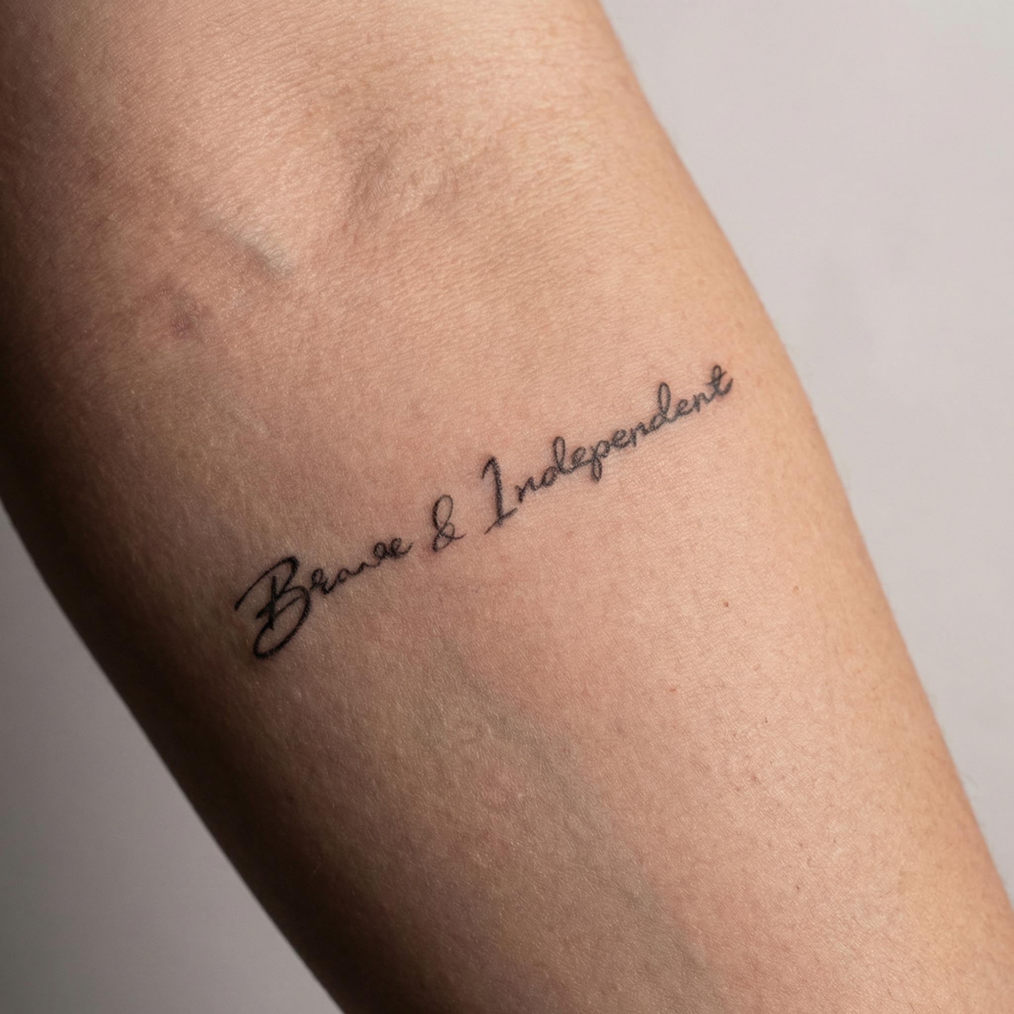 A photograph of a delicate black ink tattoo on a person's inner wrist, featuring the words "Brave & Independent" in a thin, elegant cursive font.