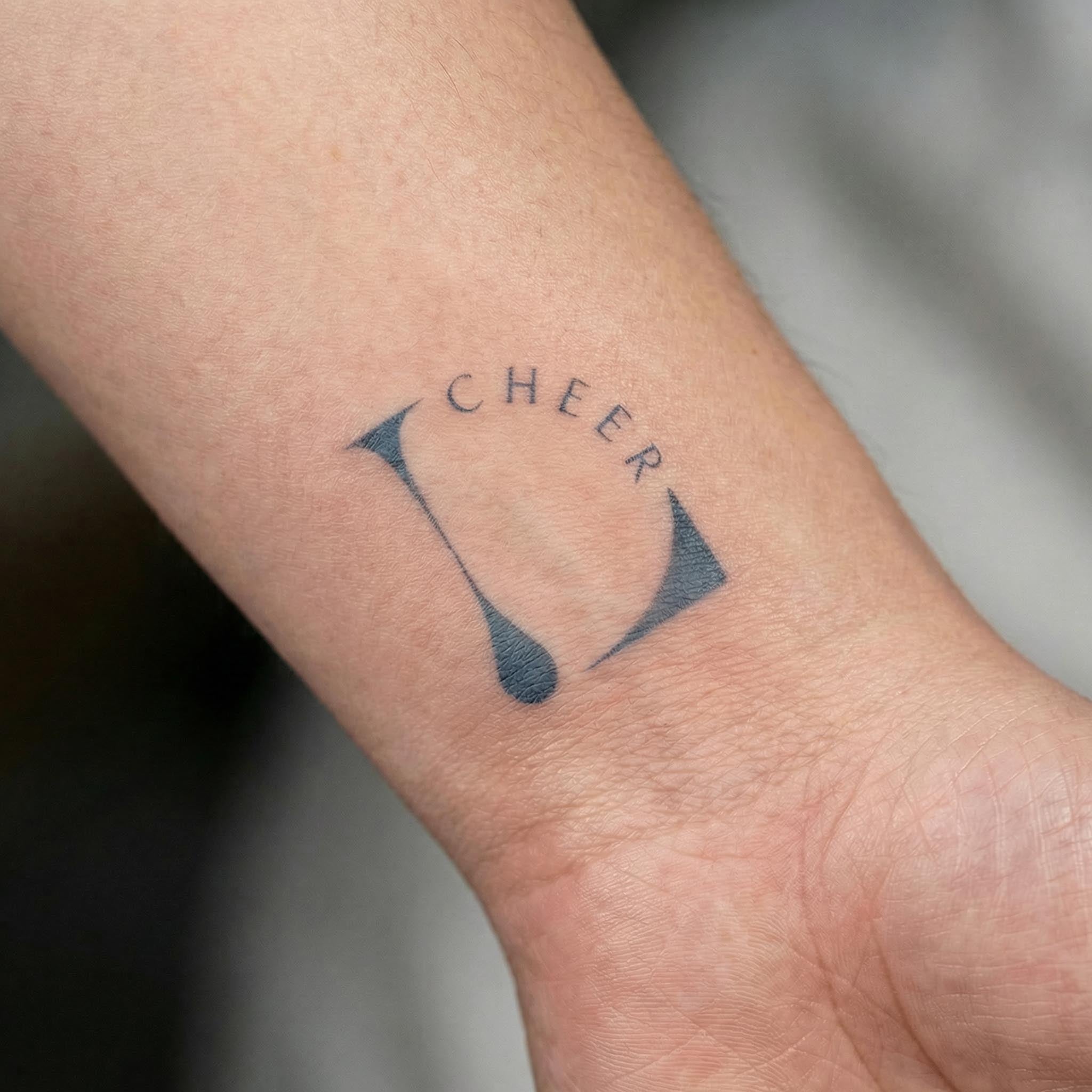 Close-up of a wrist showing a minimalist black “CHEER” temporary tattoo with curved lettering and two teardrop-shaped accents.