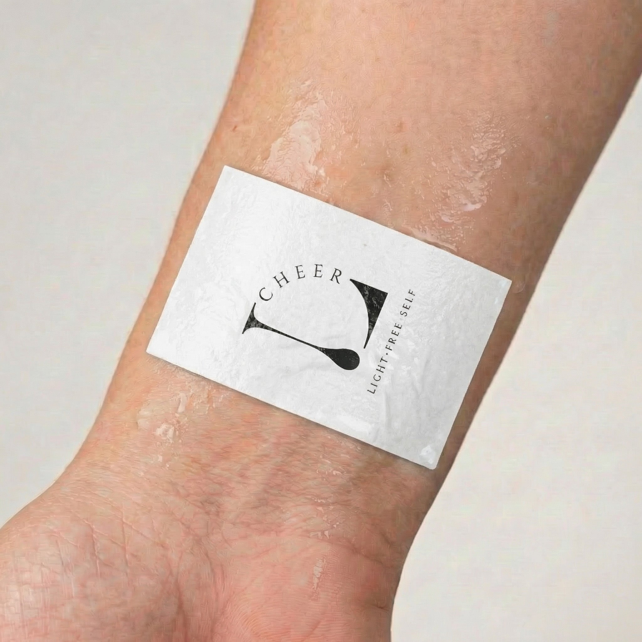 Temporary tattoo application paper on a wrist with the LCHEER logo, showing the wet transfer step before peeling.