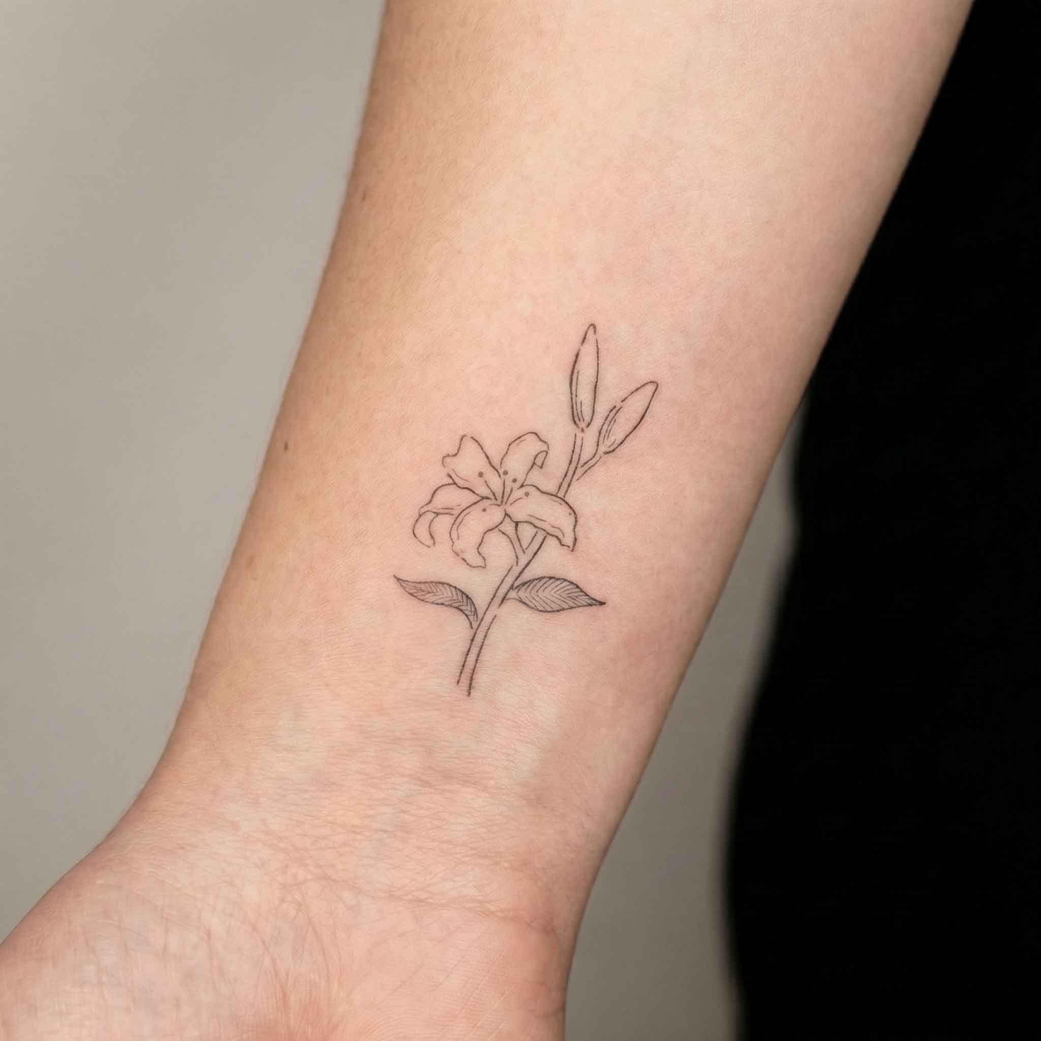 Fine line lily tattoo worn on wrist in minimalist floral style