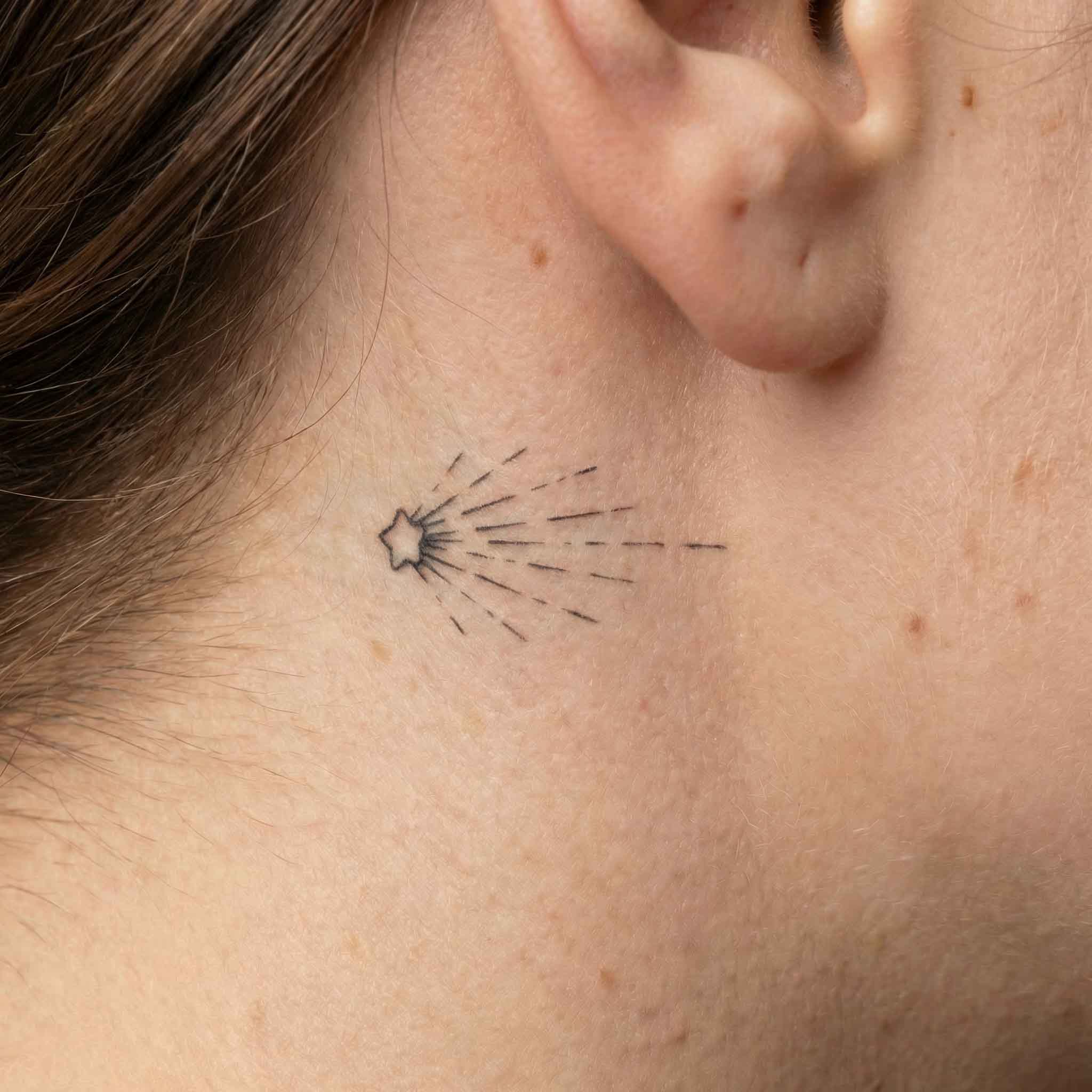 Minimal shooting star temp tattoo worn behind the ear with simple trail detail